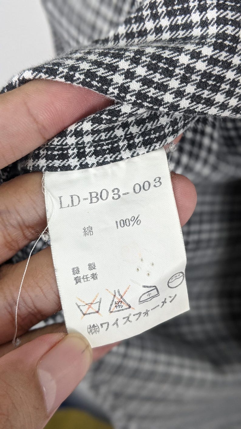 Puede incluir: Primer plano de una etiqueta de tela con el texto "LD-B03-003" y "100%" impreso. La etiqueta es blanca con texto negro y est&aacute; adherida a una tela con estampado de pata de gallo en blanco y negro. La etiqueta tambi&eacute;n incluye s&iacute;mbolos de cuidado.