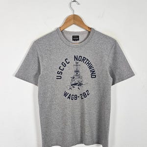 May include: A heather gray short-sleeve t-shirt featuring a dark blue graphic of a ship. The text reads USCGC NORTHWIND WAGB-282. The shirt has a crew neckline and is displayed on a wooden hanger against a plain white background.