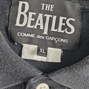 Puede incluir: Polo gris oscuro con un bot&oacute;n blanco y una etiqueta blanca con la inscripci&oacute;n "THE BEATLES COMME des GAR&Ccedil;ONS". La etiqueta tambi&eacute;n tiene una etiqueta de talla XL.