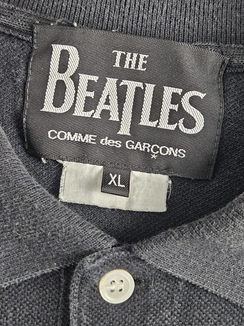 Puede incluir: Polo gris oscuro con un bot&oacute;n blanco y una etiqueta blanca con la inscripci&oacute;n "THE BEATLES COMME des GAR&Ccedil;ONS". La etiqueta tambi&eacute;n tiene una etiqueta de talla XL.