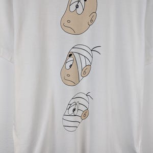 以下が含まれることがあります： さまざまな表情の3つの漫画の顔が描かれた白いTシャツ。上の顔はハゲで、真ん中の顔は包帯をしており、下の顔は包帯をしています。「COMME des GARCONS HOMME」の文字が右下にプリントされています。