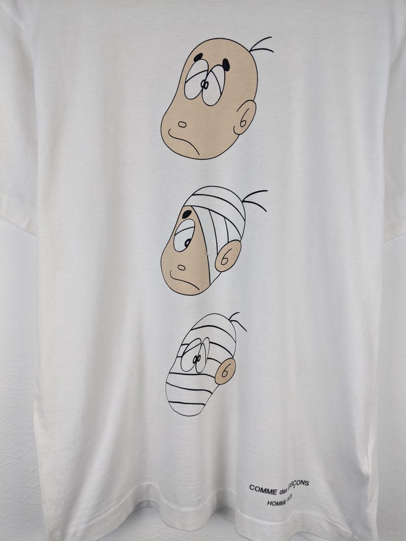 以下が含まれることがあります： さまざまな表情の3つの漫画の顔が描かれた白いTシャツ。上の顔はハゲで、真ん中の顔は包帯をしており、下の顔は包帯をしています。「COMME des GARCONS HOMME」の文字が右下にプリントされています。