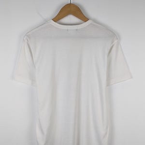 以下が含まれることがあります： 木製のハンガーに掛けられた、無地の白い半袖Tシャツ。 シャツは柔らかく軽い生地でできており、シンプルでクラシックなデザインです。 シャツは無地の白い壁を背景に表示されています。