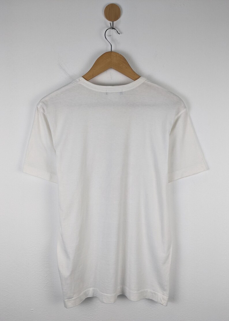 以下が含まれることがあります： 木製のハンガーに掛けられた、無地の白い半袖Tシャツ。 シャツは柔らかく軽い生地でできており、シンプルでクラシックなデザインです。 シャツは無地の白い壁を背景に表示されています。