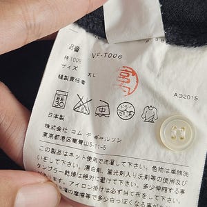Puede incluir: Primer plano de una etiqueta de ropa con texto japon&eacute;s, un bot&oacute;n y s&iacute;mbolos de cuidado. La etiqueta incluye el texto "VF-T006", "XL" y "綿100%", que indica la talla y el material de la prenda. La etiqueta tambi&eacute;n tiene un sello rojo y la fecha "AD2015".