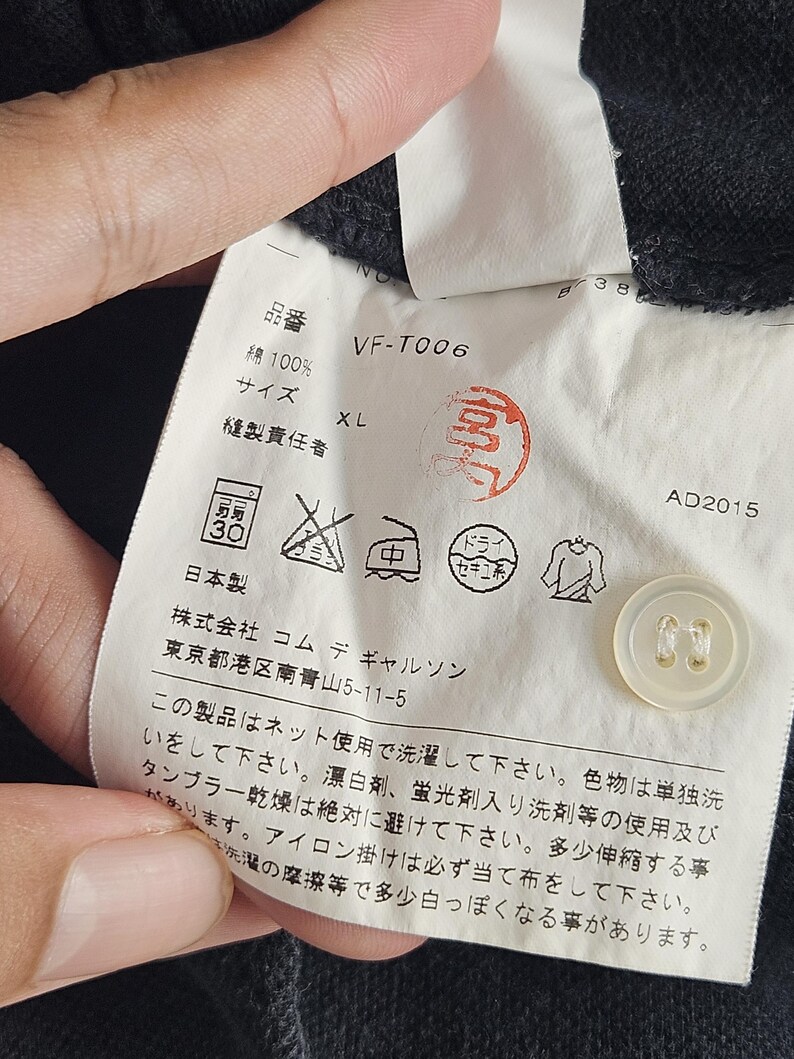 Puede incluir: Primer plano de una etiqueta de ropa con texto japon&eacute;s, un bot&oacute;n y s&iacute;mbolos de cuidado. La etiqueta incluye el texto "VF-T006", "XL" y "綿100%", que indica la talla y el material de la prenda. La etiqueta tambi&eacute;n tiene un sello rojo y la fecha "AD2015".
