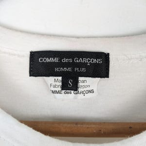 以下が含まれることがあります： 白いTシャツのクローズアップ。黒いラベルには「COMME des GARCONS HOMME PLUS」と記載されており、小さなラベルにはサイズ「S」と「Made in Japan」と記載されています。