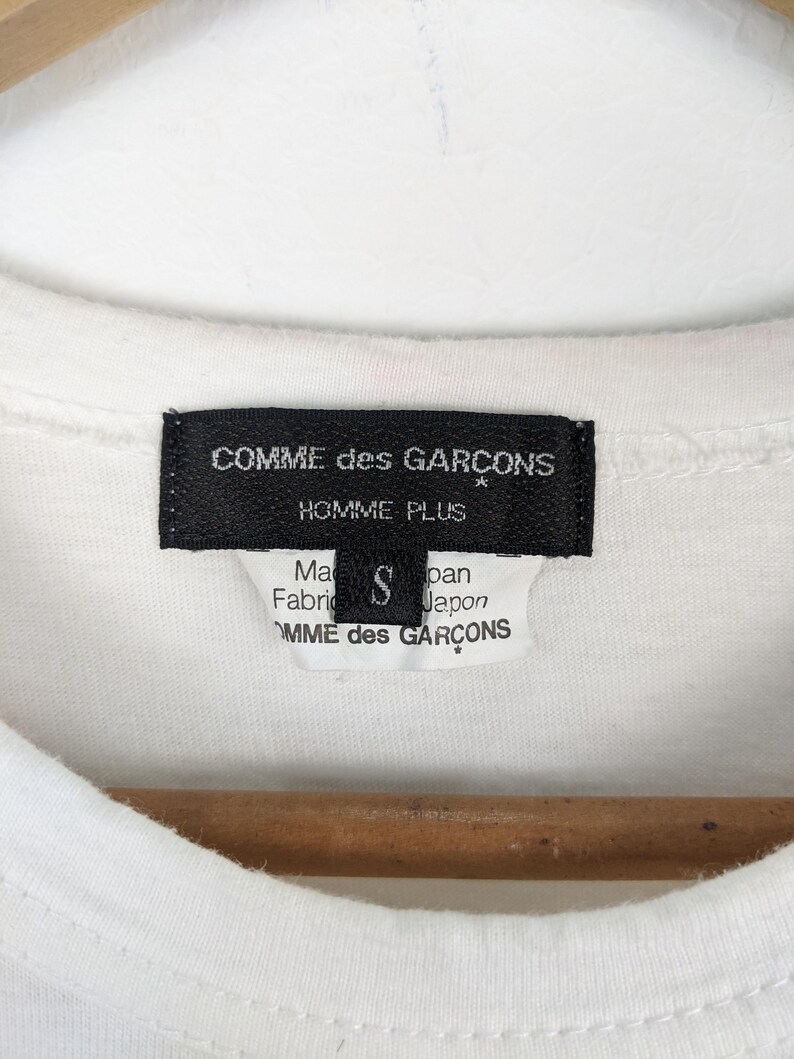 以下が含まれることがあります： 白いTシャツのクローズアップ。黒いラベルには「COMME des GARCONS HOMME PLUS」と記載されており、小さなラベルにはサイズ「S」と「Made in Japan」と記載されています。