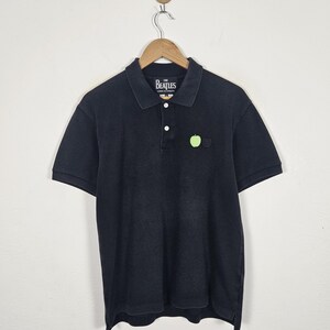 Puede incluir: Polo negro con mangas cortas, cuello y tapeta de tres botones. La camisa presenta un peque&ntilde;o emblema de manzana verde y una etiqueta que dice "The Beatles". La camisa est&aacute; colgada de una percha de madera.