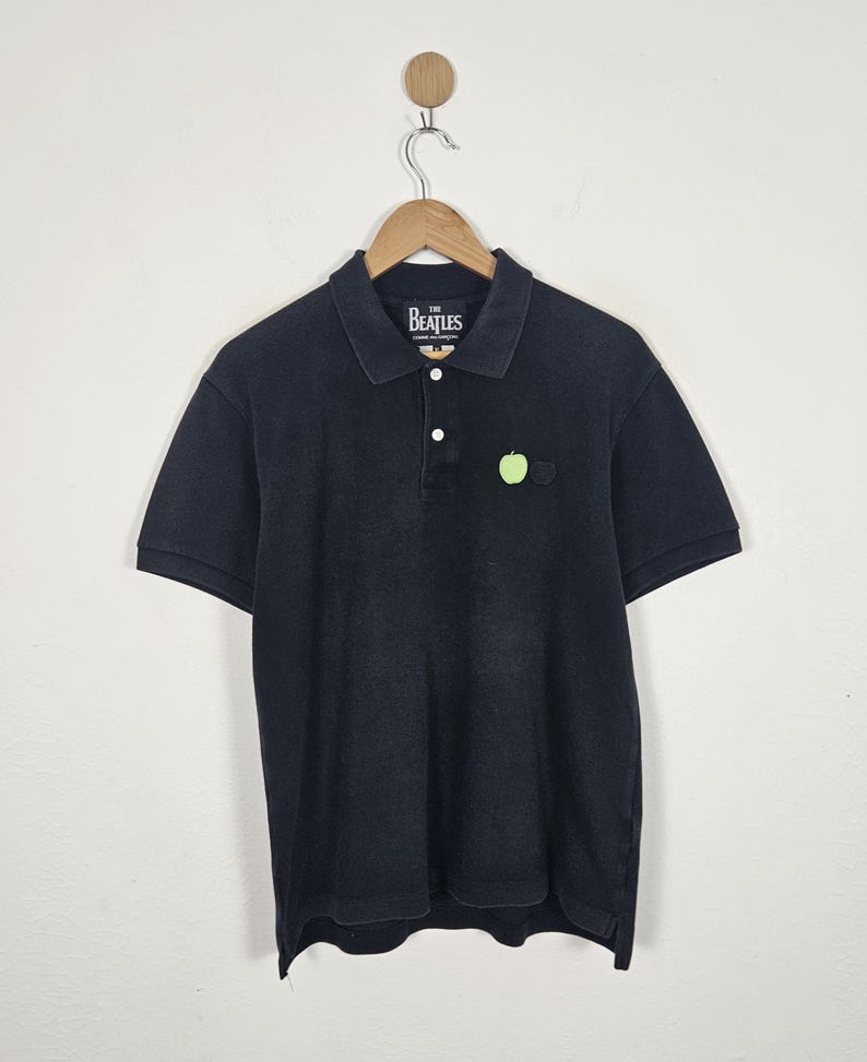 Puede incluir: Polo negro con mangas cortas, cuello y tapeta de tres botones. La camisa presenta un peque&ntilde;o emblema de manzana verde y una etiqueta que dice "The Beatles". La camisa est&aacute; colgada de una percha de madera.