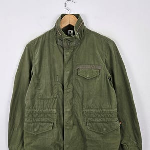 May include: An olive green jacket with a stand-up collar, multiple pockets, and a button-up front. The jacket is hanging on a wooden hanger against a white background. The jacket has a military-inspired design.