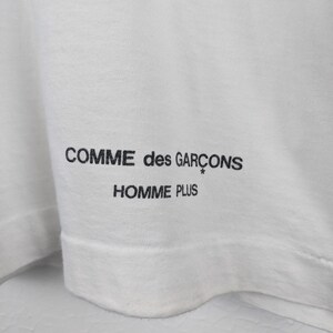 以下が含まれることがあります： 白のTシャツのクローズアップ。黒色の「COMME des GAR&Ccedil;ONS HOMME PLUS」のテキストがプリントされています。テキストはシャツの裾の近くにあります。生地は柔らかく、軽量な素材のようです。