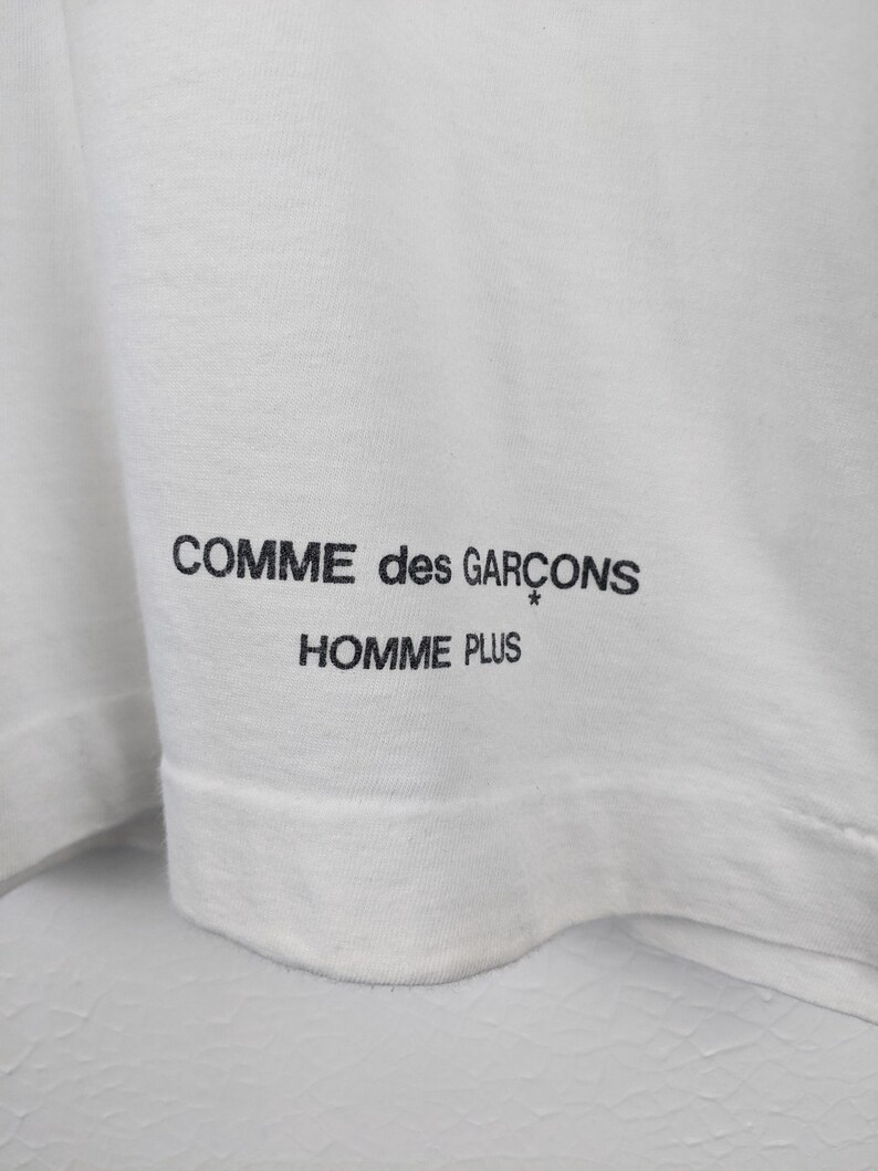 以下が含まれることがあります： 白のTシャツのクローズアップ。黒色の「COMME des GAR&Ccedil;ONS HOMME PLUS」のテキストがプリントされています。テキストはシャツの裾の近くにあります。生地は柔らかく、軽量な素材のようです。
