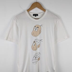 以下が含まれることがあります： 3つの漫画の顔が描かれた白いTシャツ。それぞれの顔は異なる表情と包帯をしています。Tシャツはクルーネックと半袖です。ブランド名はTシャツの上部と下部に表示されています。