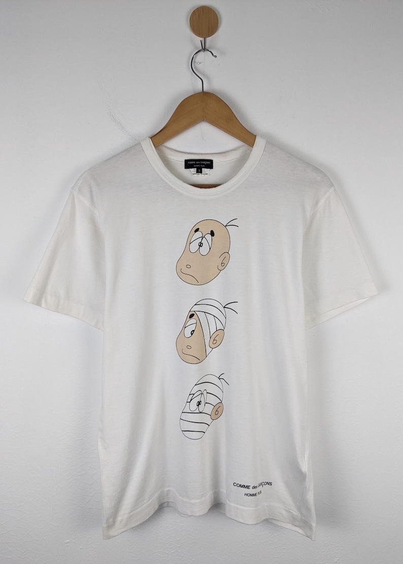 以下が含まれることがあります： 3つの漫画の顔が描かれた白いTシャツ。それぞれの顔は異なる表情と包帯をしています。Tシャツはクルーネックと半袖です。ブランド名はTシャツの上部と下部に表示されています。