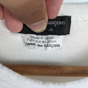 以下が含まれることがあります： 白いTシャツのクローズアップ。黒いタグには「COMME des GARCONS」と「Made in Japan / Fabriqu&eacute; au Japon」と書かれています。Tシャツは木製のハンガーにかかっています。
