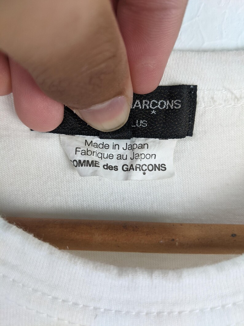 以下が含まれることがあります： 白いTシャツのクローズアップ。黒いタグには「COMME des GARCONS」と「Made in Japan / Fabriqu&eacute; au Japon」と書かれています。Tシャツは木製のハンガーにかかっています。