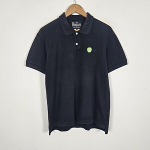 Puede incluir: Polo negro de manga corta, con cuello y tapeta abotonada. La camisa tiene un peque&ntilde;o logotipo de manzana verde y el texto "The Beatles" en el cuello. Cuelga de una percha de madera.