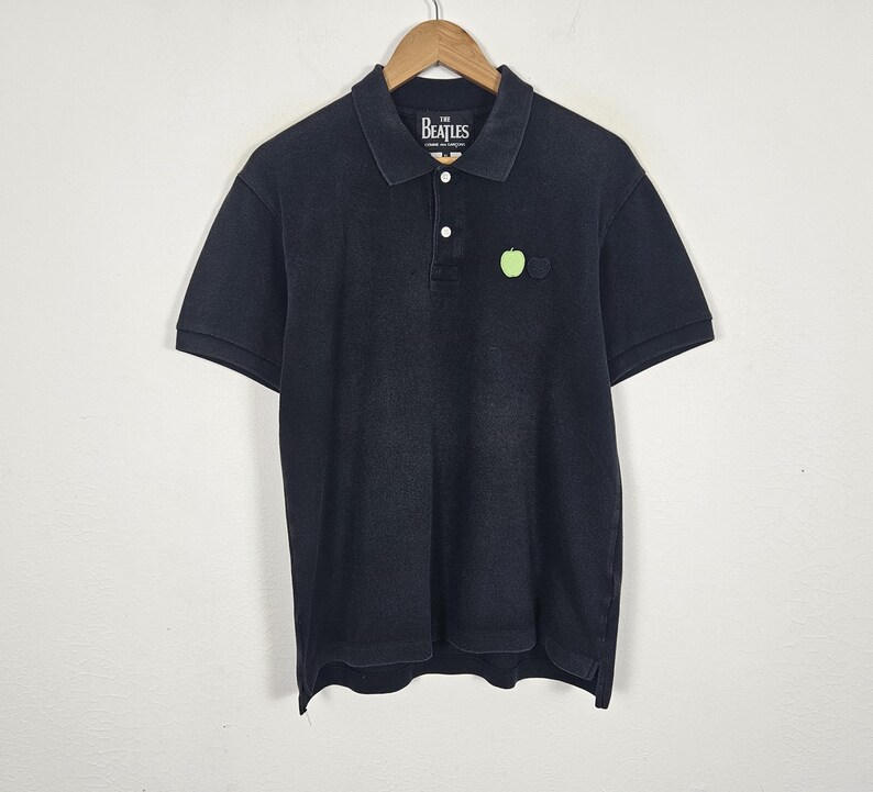 Puede incluir: Polo negro de manga corta, con cuello y tapeta abotonada. La camisa tiene un peque&ntilde;o logotipo de manzana verde y el texto "The Beatles" en el cuello. Cuelga de una percha de madera.