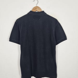Puede incluir: Polo negro de manga corta, colgado de una percha de madera. La camisa tiene un cuello cl&aacute;sico y un dise&ntilde;o sencillo. La tela parece ser suave y transpirable, adecuada para ropa informal.