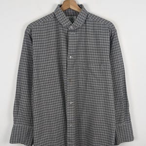 Puede incluir: Camisa de manga larga con estampado de cuadros blancos y negros. La camisa tiene un bolsillo en el pecho y un cuello cl&aacute;sico. La prenda est&aacute; colgada de una percha de madera sobre un fondo blanco.