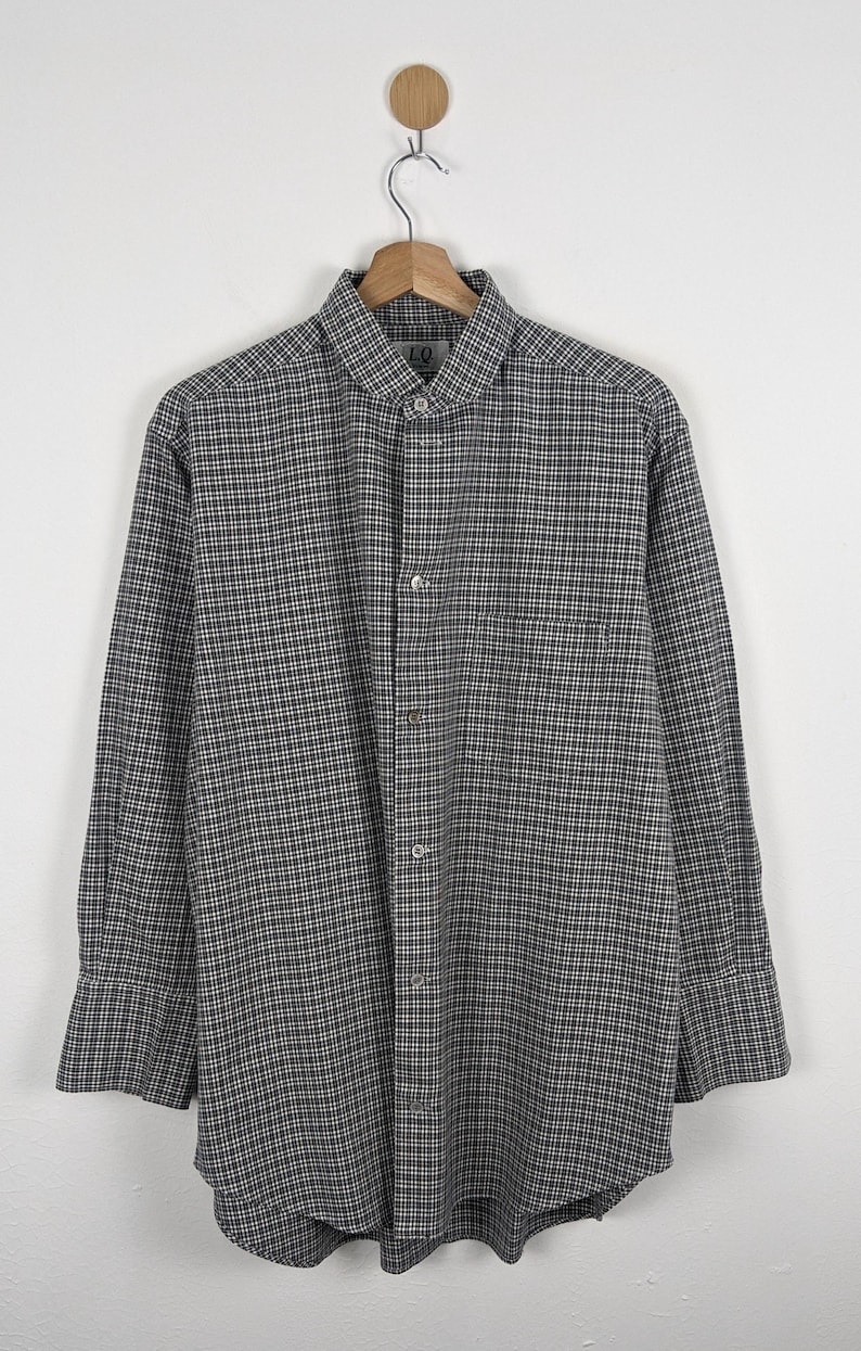 Puede incluir: Camisa de manga larga con estampado de cuadros blancos y negros. La camisa tiene un bolsillo en el pecho y un cuello cl&aacute;sico. La prenda est&aacute; colgada de una percha de madera sobre un fondo blanco.