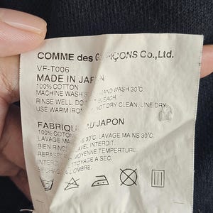 Puede incluir: Una etiqueta blanca con texto negro de Comme des Gar&ccedil;ons Co., Ltd. dice "MADE IN JAPAN" e instrucciones de cuidado. La etiqueta est&aacute; hecha de 100% algod&oacute;n e incluye s&iacute;mbolos de lavado y secado. La etiqueta est&aacute; sujeta por una mano contra una tela oscura.