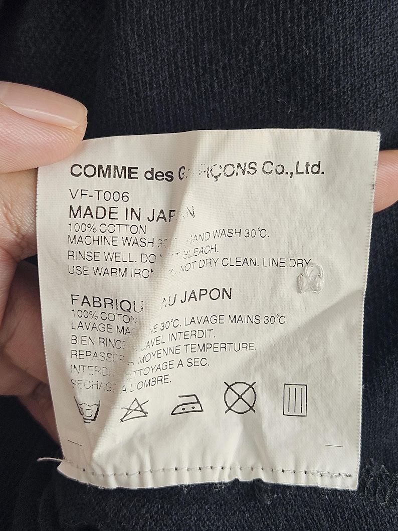 Puede incluir: Una etiqueta blanca con texto negro de Comme des Gar&ccedil;ons Co., Ltd. dice "MADE IN JAPAN" e instrucciones de cuidado. La etiqueta est&aacute; hecha de 100% algod&oacute;n e incluye s&iacute;mbolos de lavado y secado. La etiqueta est&aacute; sujeta por una mano contra una tela oscura.