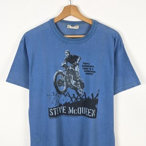 May include: Blue t-shirt featuring a black and white graphic of Steve McQueen riding a motorcycle. The shirt includes the text "STEVE MCQUEEN" and a quote about barbed wire.