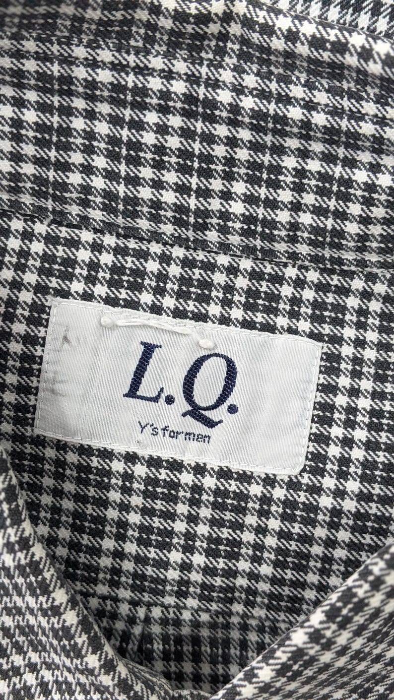 Puede incluir: Primer plano de una camisa con estampado de pata de gallo en blanco y negro. La camisa tiene una etiqueta que dice "L.Q. Y's for men". La tela est&aacute; tejida de forma apretada, creando una apariencia texturizada. La est&eacute;tica general es cl&aacute;sica y vers&aacute;til.
