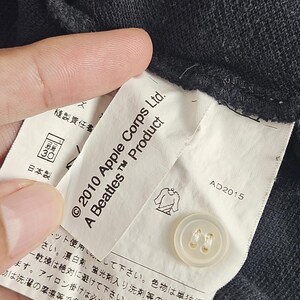 Puede incluir: Primer plano de una etiqueta de prenda gris oscuro con el texto "&copy; 2010 Apple Corps Ltd. A Beatles Product". Se ve un bot&oacute;n blanco. La etiqueta tambi&eacute;n tiene texto japon&eacute;s y una ilustraci&oacute;n de camisa.