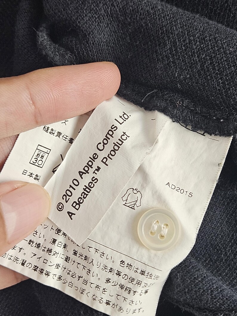 Puede incluir: Primer plano de una etiqueta de prenda gris oscuro con el texto "&copy; 2010 Apple Corps Ltd. A Beatles Product". Se ve un bot&oacute;n blanco. La etiqueta tambi&eacute;n tiene texto japon&eacute;s y una ilustraci&oacute;n de camisa.
