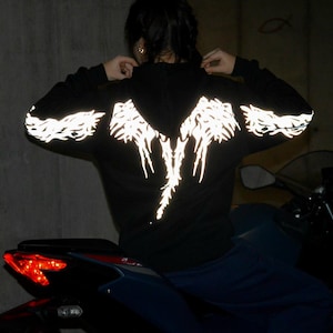May include: A black hooded sweatshirt featuring a reflective silver graphic design on the back and sleeves. The design consists of intricate, wing-like patterns that illuminate in low light conditions, extending across the shoulders and down the arms.