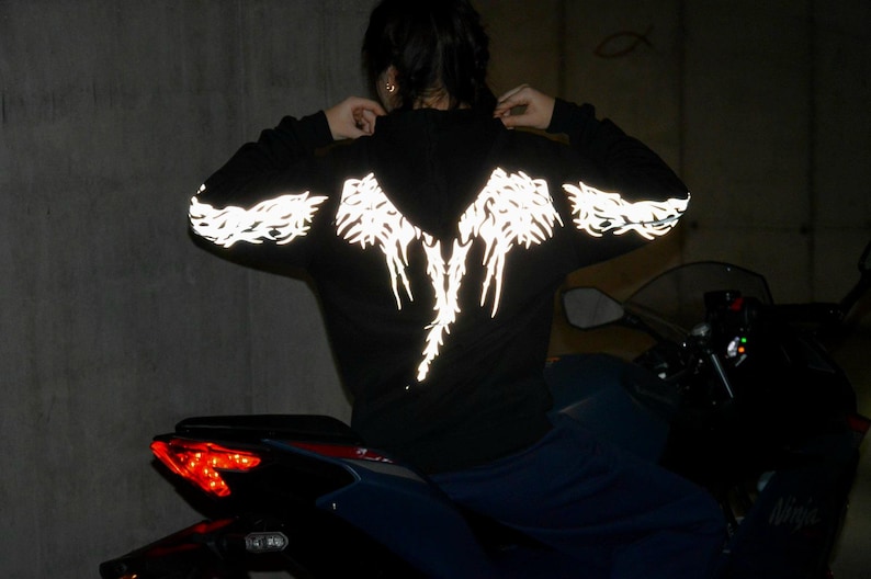 May include: A black hooded sweatshirt featuring a reflective silver graphic design on the back and sleeves. The design consists of intricate, wing-like patterns that illuminate in low light conditions, extending across the shoulders and down the arms.