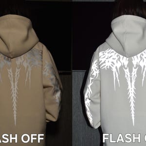 May include: A beige hooded sweatshirt featuring a reflective tribal-style graphic pattern across the back and sleeves. The image displays two views: one labeled FLASH OFF showing the beige fabric, and one labeled FLASH ON showing the reflective design.