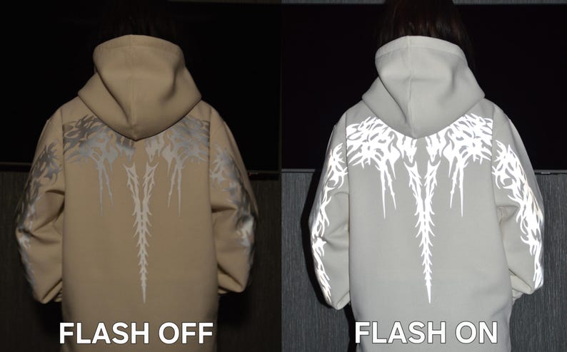 May include: A beige hooded sweatshirt featuring a reflective tribal-style graphic pattern across the back and sleeves. The image displays two views: one labeled FLASH OFF showing the beige fabric, and one labeled FLASH ON showing the reflective design.