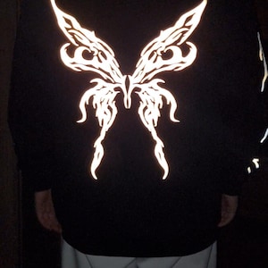 Puede incluir: Sudadera con capucha negra con un gran dise&ntilde;o reflectante de alas de mariposa en la espalda. Las alas son detalladas y brillan. La sudadera se combina con pantalones de color claro.