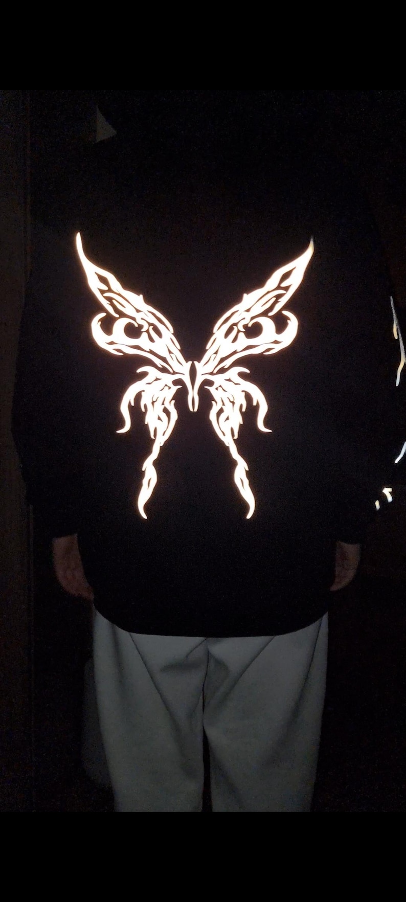 Puede incluir: Sudadera con capucha negra con un gran dise&ntilde;o reflectante de alas de mariposa en la espalda. Las alas son detalladas y brillan. La sudadera se combina con pantalones de color claro.