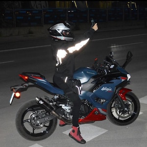 May include: A blue and red Kawasaki Ninja motorcycle with black wheels and a black exhaust pipe. The rider wears a black helmet, a black hooded sweatshirt with reflective white sleeve details, black pants, and red and black high-top sneakers.