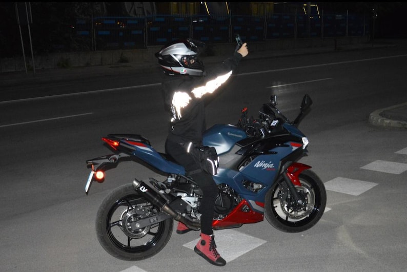 May include: A blue and red Kawasaki Ninja motorcycle with black wheels and a black exhaust pipe. The rider wears a black helmet, a black hooded sweatshirt with reflective white sleeve details, black pants, and red and black high-top sneakers.