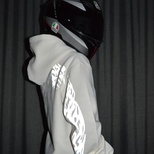 May include: A white hooded sweatshirt featuring reflective flame-like patterns along the sleeves, paired with a grey and black full-face motorcycle helmet displaying the AGV logo on the side, captured from a side profile perspective.