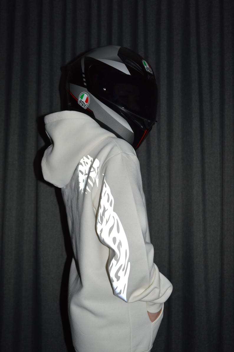 May include: A white hooded sweatshirt featuring reflective flame-like patterns along the sleeves, paired with a grey and black full-face motorcycle helmet displaying the AGV logo on the side, captured from a side profile perspective.