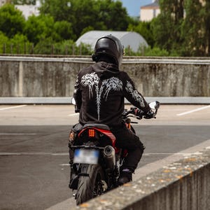 May include: A black hooded sweatshirt featuring a white graphic design on the back and sleeves, worn by a rider on a red motorcycle. The rider wears a black helmet and black pants. The motorcycle has a visible rear tire, exhaust, and red taillight.