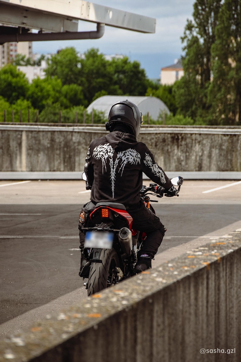 May include: A black hooded sweatshirt featuring a white graphic design on the back and sleeves, worn by a rider on a red motorcycle. The rider wears a black helmet and black pants. The motorcycle has a visible rear tire, exhaust, and red taillight.