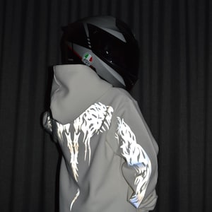 May include: A light beige hooded sweatshirt featuring reflective silver tribal-style patterns on the back and sleeves. The garment is worn with a black and grey full-face motorcycle helmet displaying an AGV logo on the side.