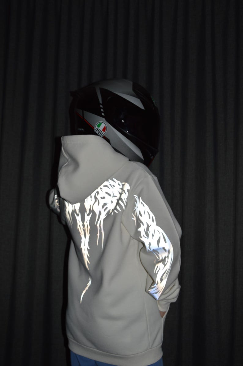 May include: A light beige hooded sweatshirt featuring reflective silver tribal-style patterns on the back and sleeves. The garment is worn with a black and grey full-face motorcycle helmet displaying an AGV logo on the side.