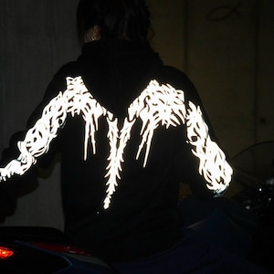 May include: A black hooded sweatshirt featuring a reflective white graphic pattern that spans across the back and down both sleeves. The design consists of intricate, sharp, flame-like lines that illuminate brightly against the dark fabric.