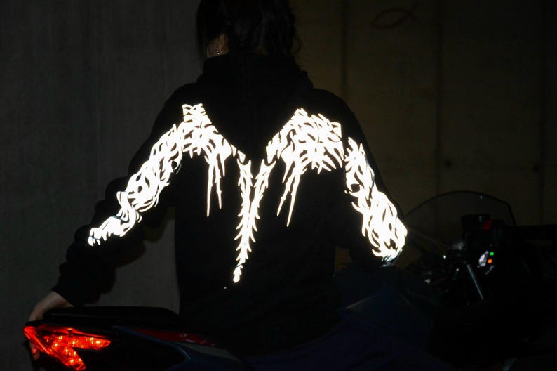 May include: A black hooded sweatshirt featuring a reflective white graphic pattern that spans across the back and down both sleeves. The design consists of intricate, sharp, flame-like lines that illuminate brightly against the dark fabric.