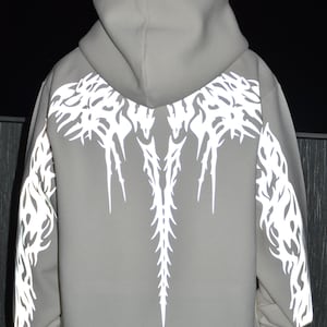 May include: A light gray hooded sweatshirt featuring a reflective white graphic print on the back and sleeves. The design consists of intricate, sharp, flame-like patterns that extend from the shoulders down the back and along the outer arms.