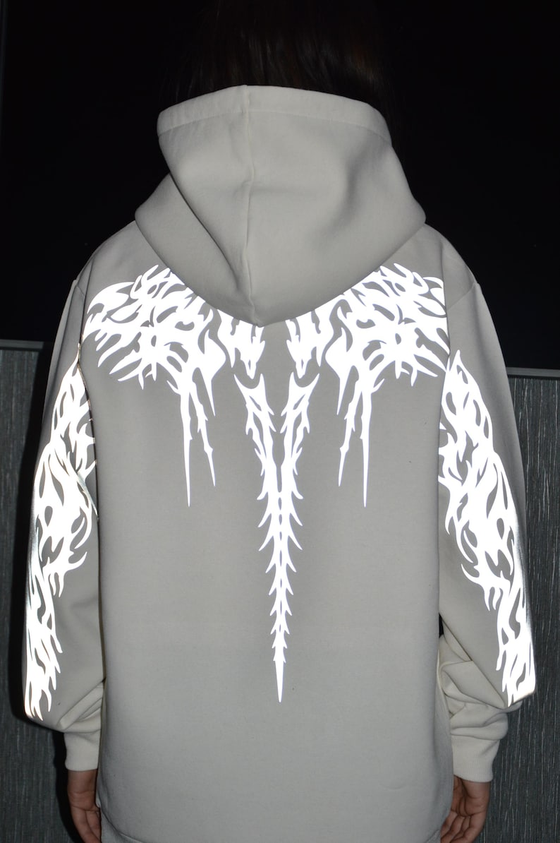 May include: A light gray hooded sweatshirt featuring a reflective white graphic print on the back and sleeves. The design consists of intricate, sharp, flame-like patterns that extend from the shoulders down the back and along the outer arms.